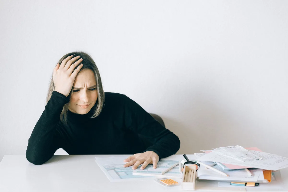 Person overwhelmed by a pile of paperwork at a desk, looking discouraged