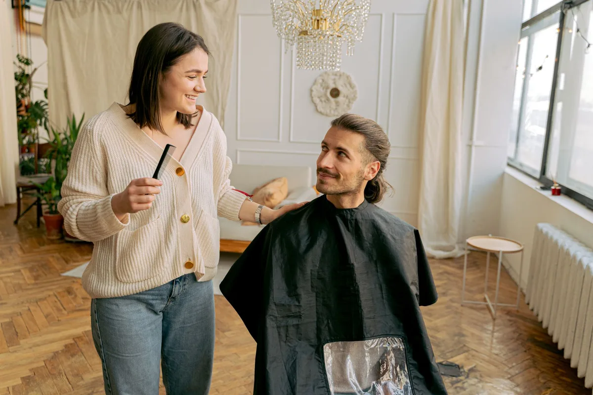 Coiffeuse souriante, peigne à la main, qui discute avec son client assis sous la cape dans un salon lumineux, un échange qui commence par la langue du client