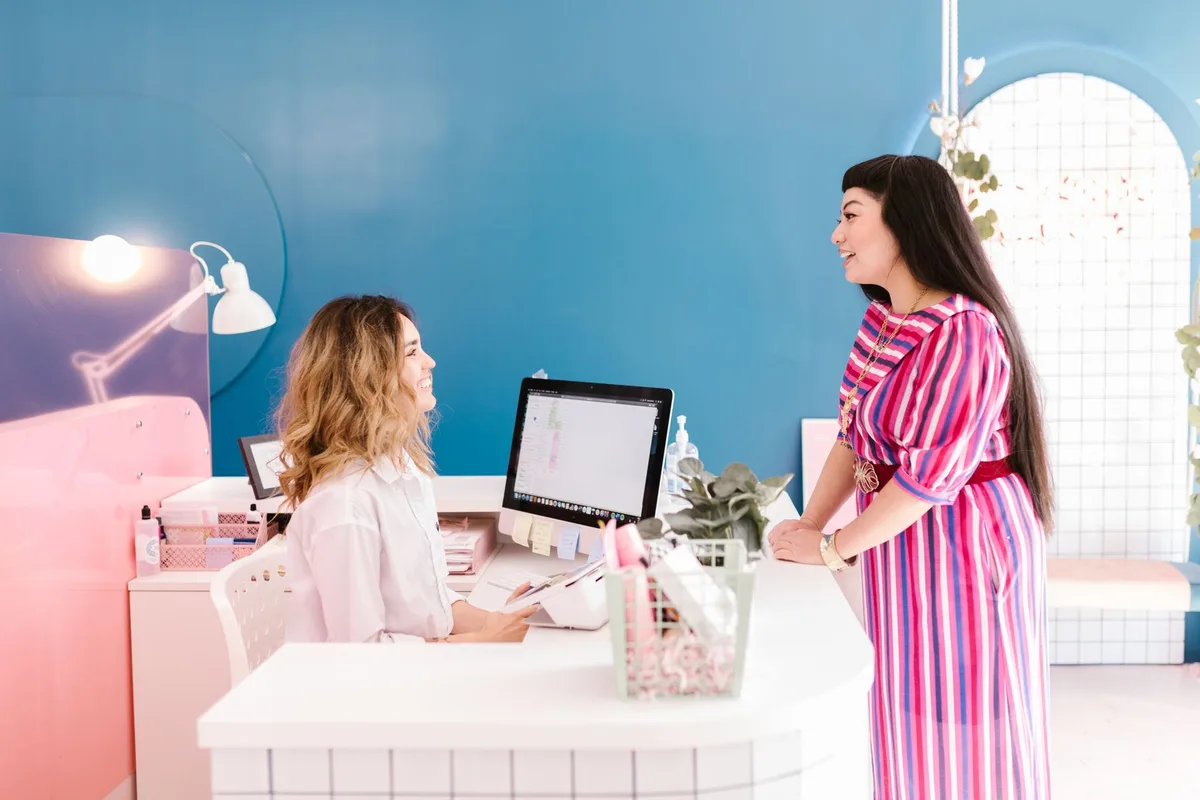 Salon receptionist at her calendar screen, chatting with a client at the front desk, like the moment the next visit gets booked to retain a new client