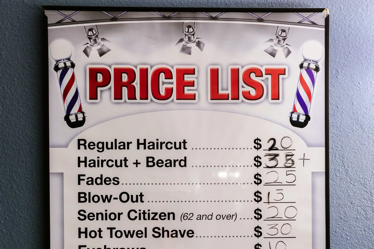 Affiche de tarifs encadrée au mur d'un barbershop, aux noms de services abrégés et aux prix corrigés à la main